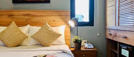 Un dormitorio con cabecera de madera, cama con ropa blanca, mesita de noche con lámpara, ventana y armario de madera.
