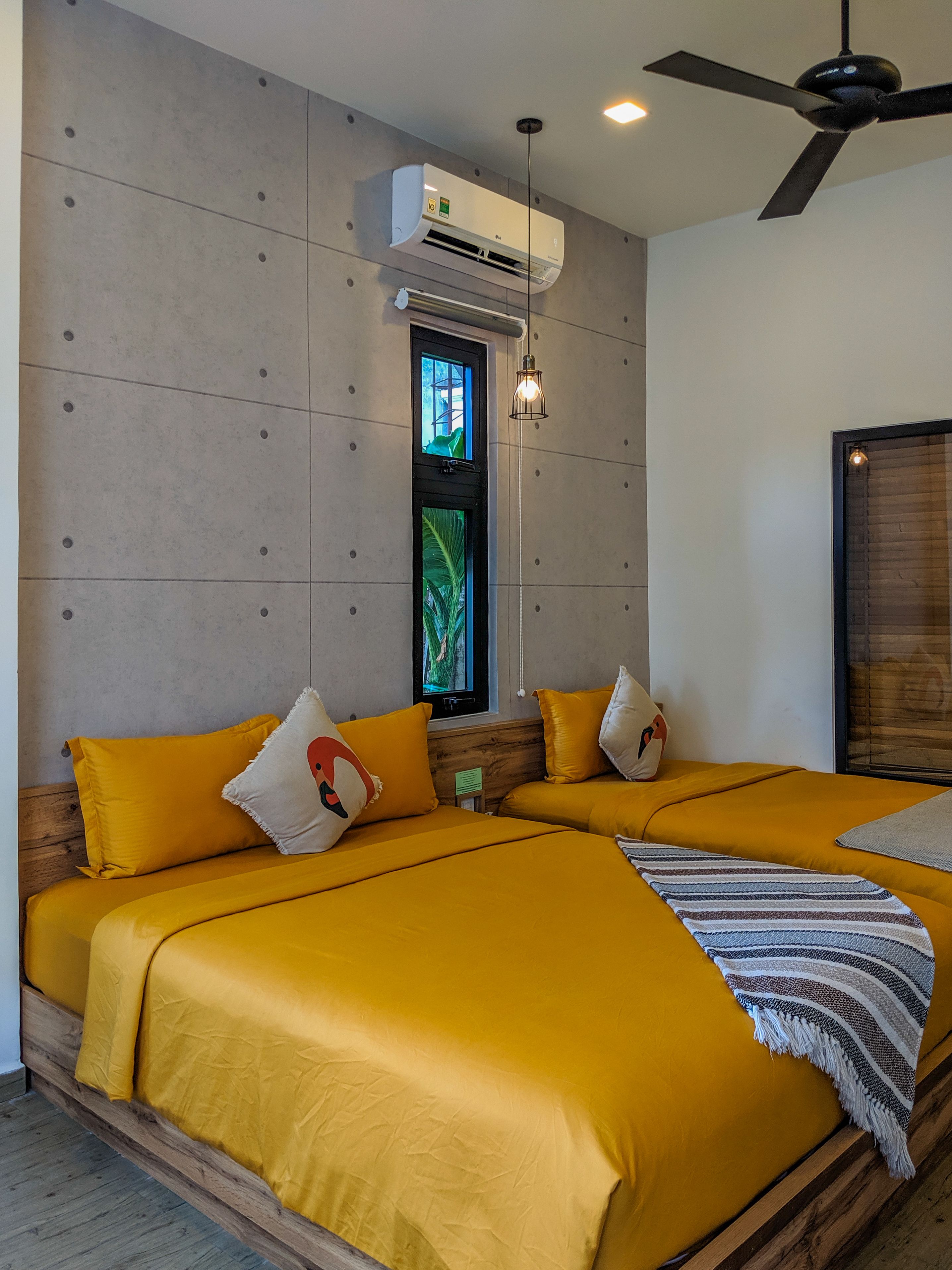 A room with two beds, each with yellow bedding and decorative pillows, a ceiling fan, an air conditioning unit, and a window with a view of greenery.