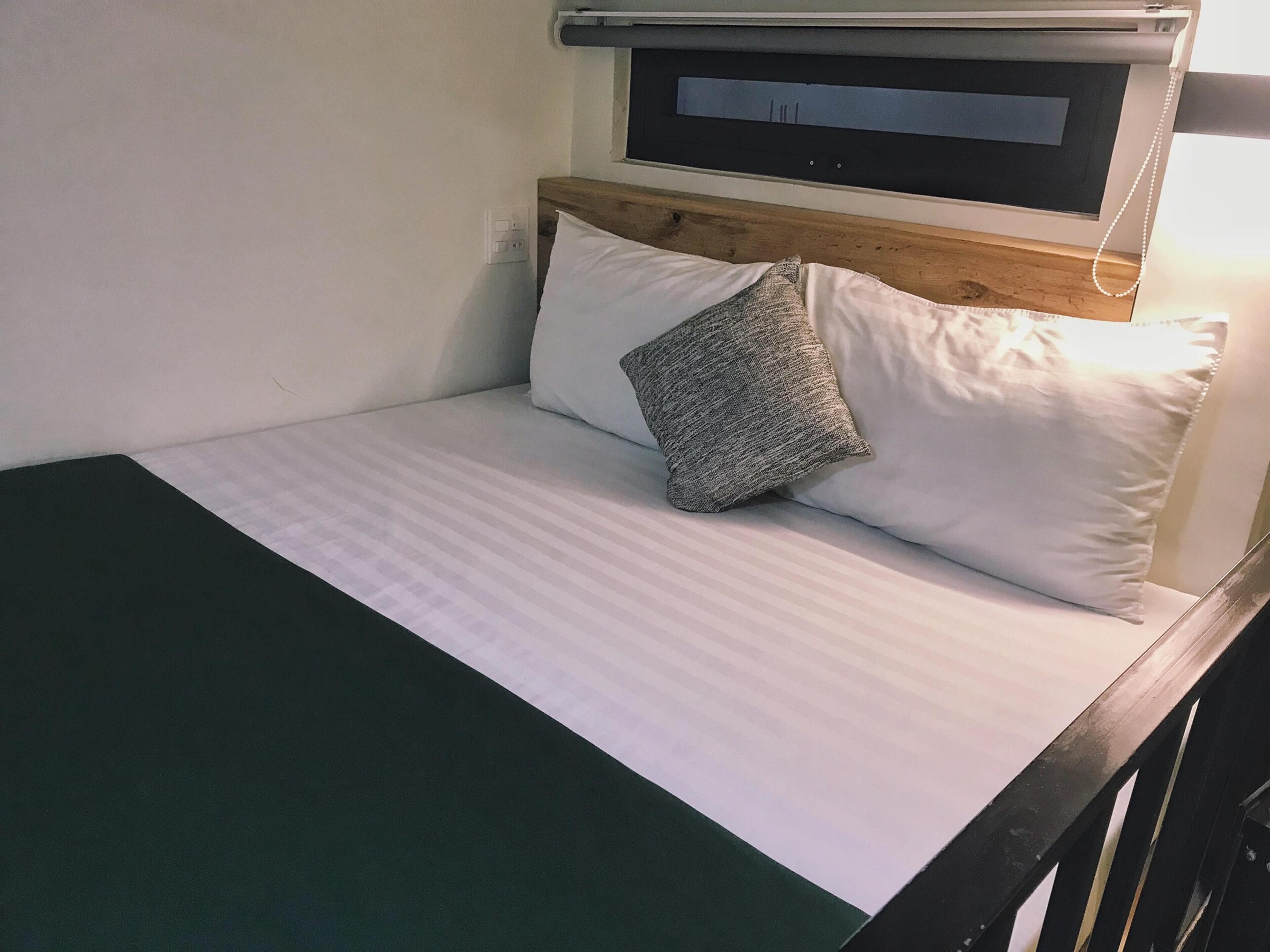 A bed with white bedding and a grey pillow, a wooden headboard, and a small window above the bed.