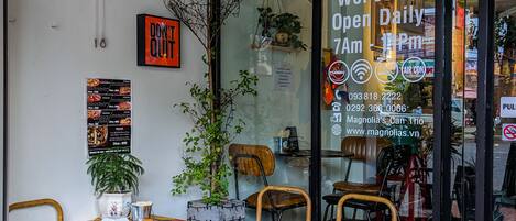 An interior with a wooden table and chairs, a potted plant, and a glass door with a sign indicating the cafe is open daily from 7 AM to 11 PM.