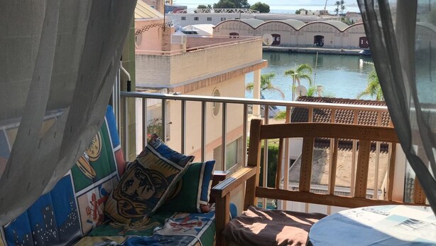 Ein Balkon mit einem Holzstuhl und einem farbenfrohen Sofa, mit Blick auf einen Yachthafen und Gebäude.