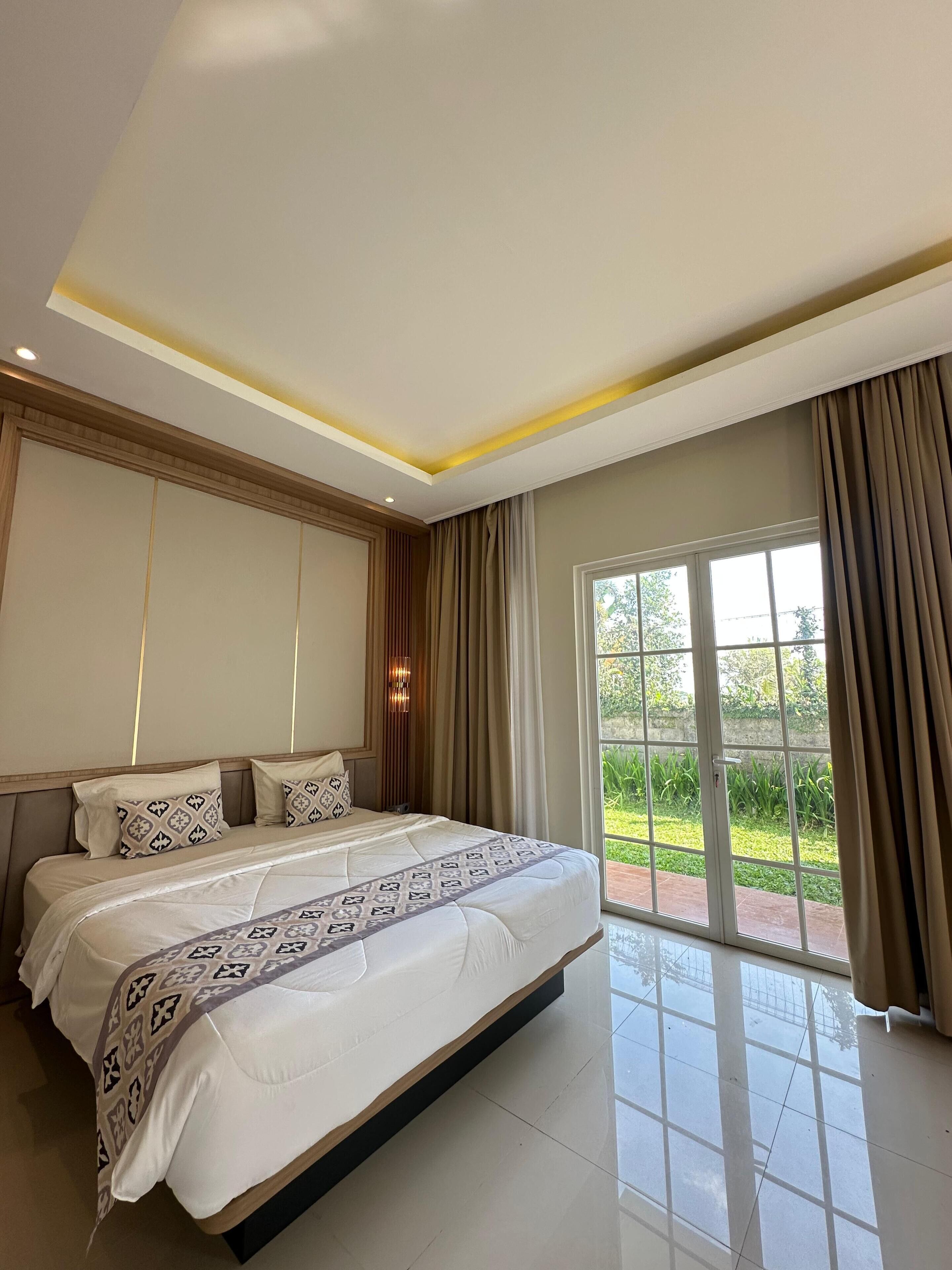 A modern bedroom with a large bed, a view of greenery through the window, and a ceiling with recessed lighting.
