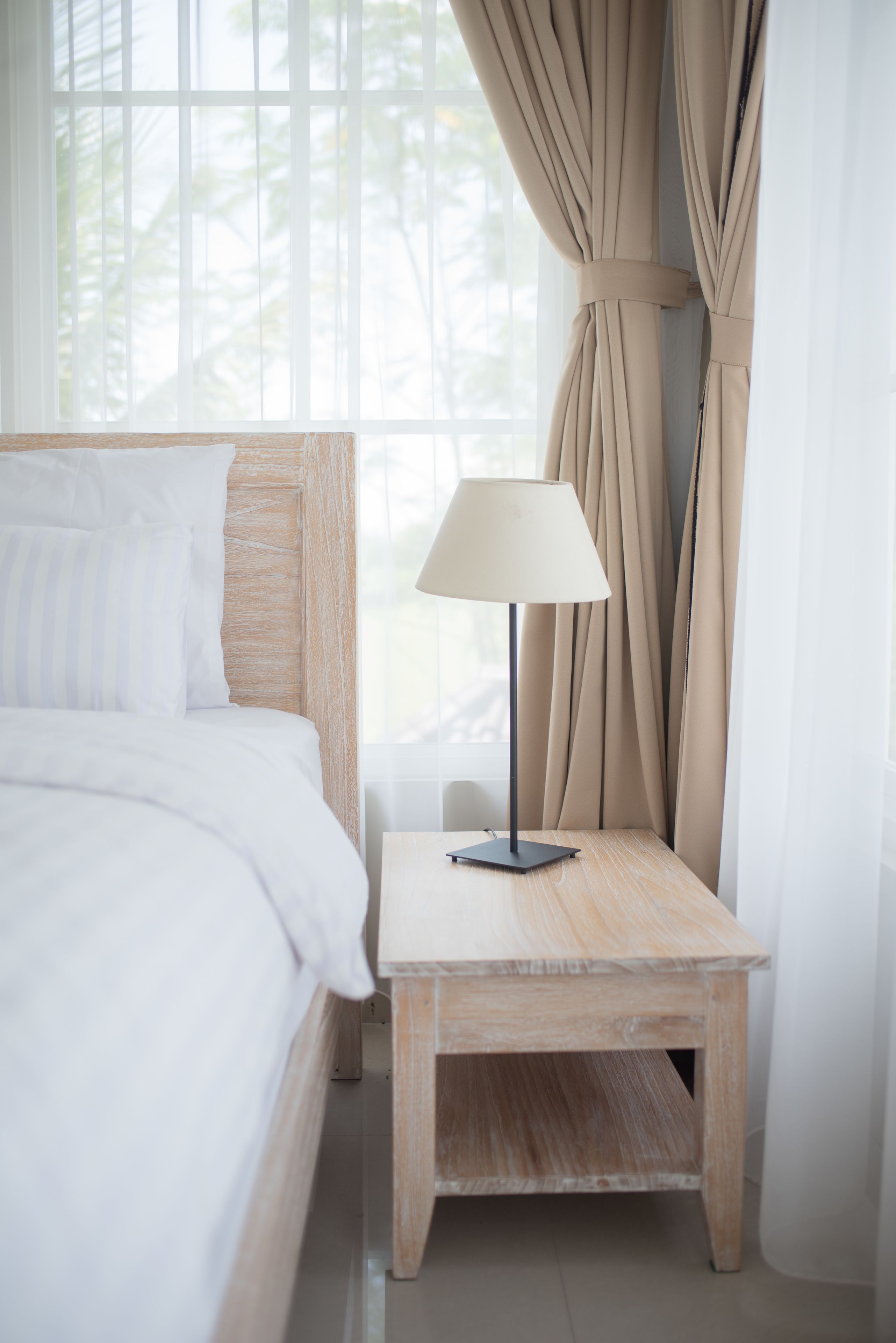 A neatly made bed with white bedding, a bedside table with a lamp, and beige curtains.