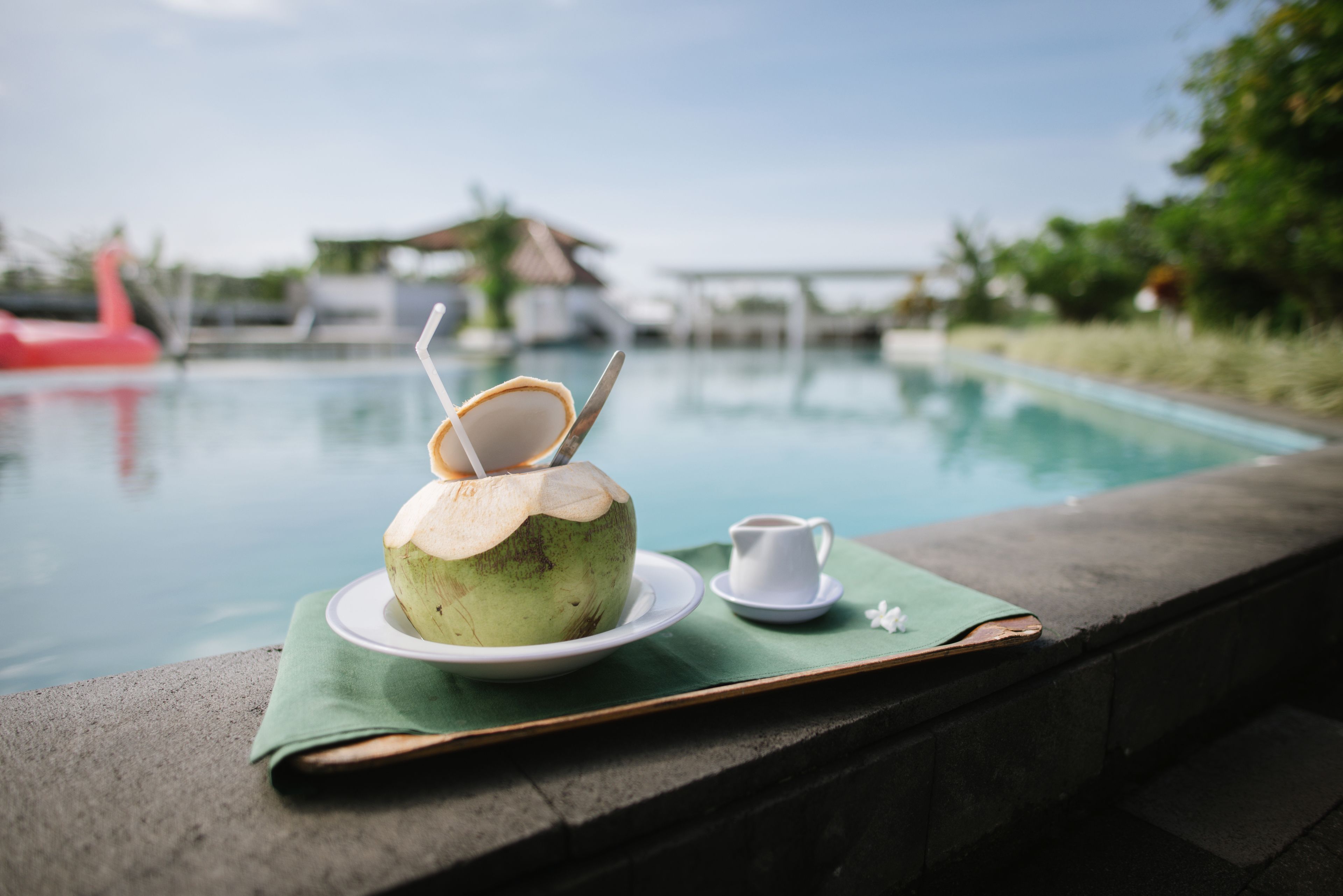 A coconut drink with a straw and a small cup on a green mat by a poolside.