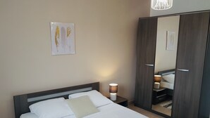 A neatly made bed with white linens and a beige pillow, a bedside table with a lamp, a large wardrobe, and a framed artwork on the wall.