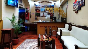 Zona de recepción con un sofá, una silla de madera, mostrador de recepción, televisión y una pared con el letrero 'WELCOME'.