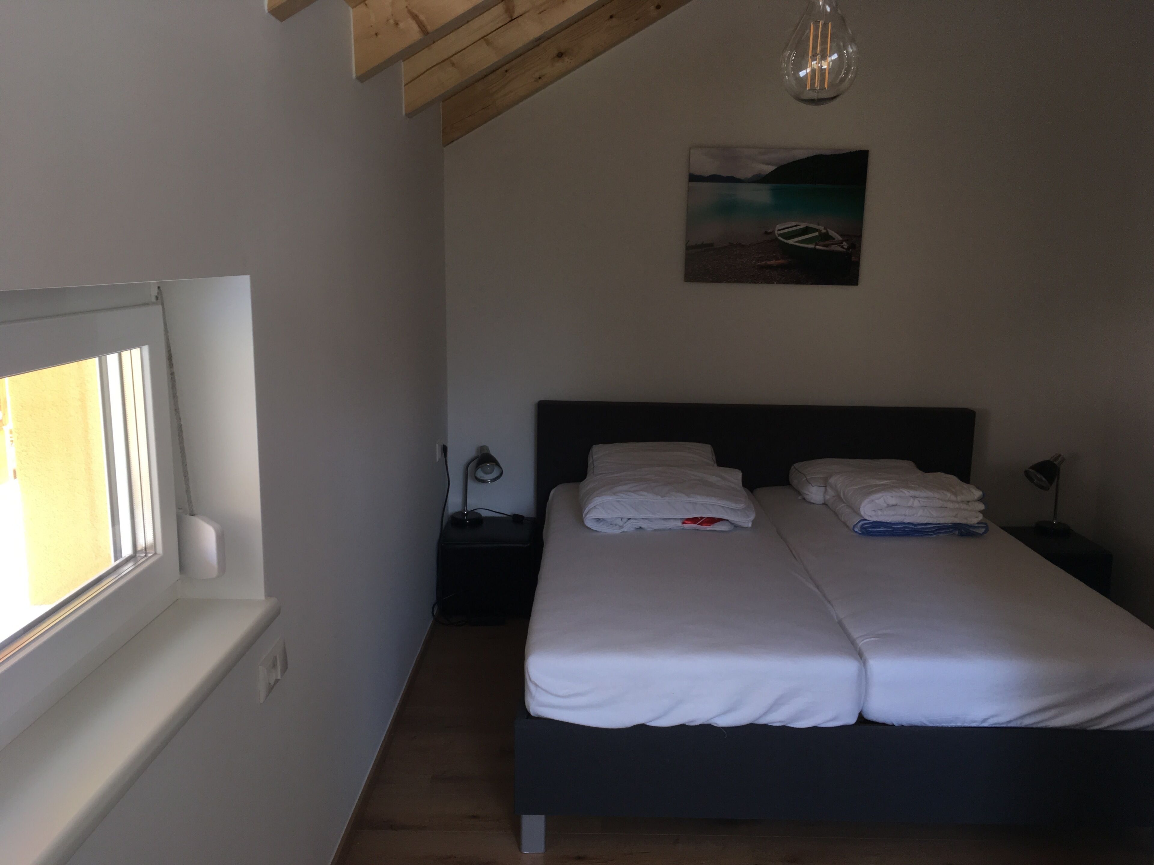 A bedroom with a double bed, a nightstand with a lamp, a painting on the wall, and a window with blinds.