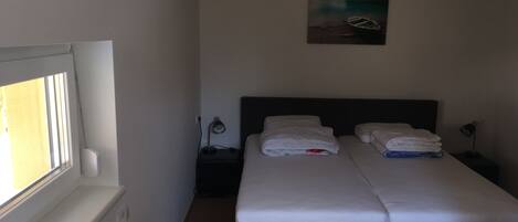 A bedroom with a double bed, a nightstand with a lamp, a painting on the wall, and a window with blinds.