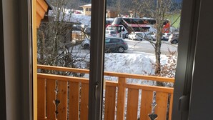 Blick von einem Balkon mit Holzgeländer auf einen Parkplatz mit Autos und einem Bus, im Hintergrund eine Berglandschaft.