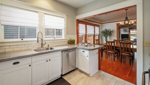 A kitchen with white cabinets, a dishwasher, and a dining area with wooden chairs and a table. - Historic 1910 Bungalow & Carriage House • Dog Friendly • 1 Block to Wine & Dine (McMinnville)