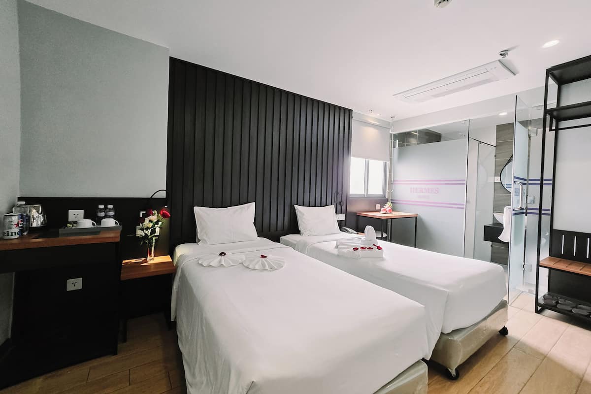 A hotel room with two beds, a desk with a computer, a small table with a vase of flowers, and a glass-enclosed shower area.