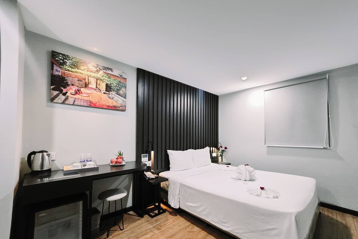 A hotel room with a bed, a desk with a chair, a microwave, a kettle, and a painting on the wall.