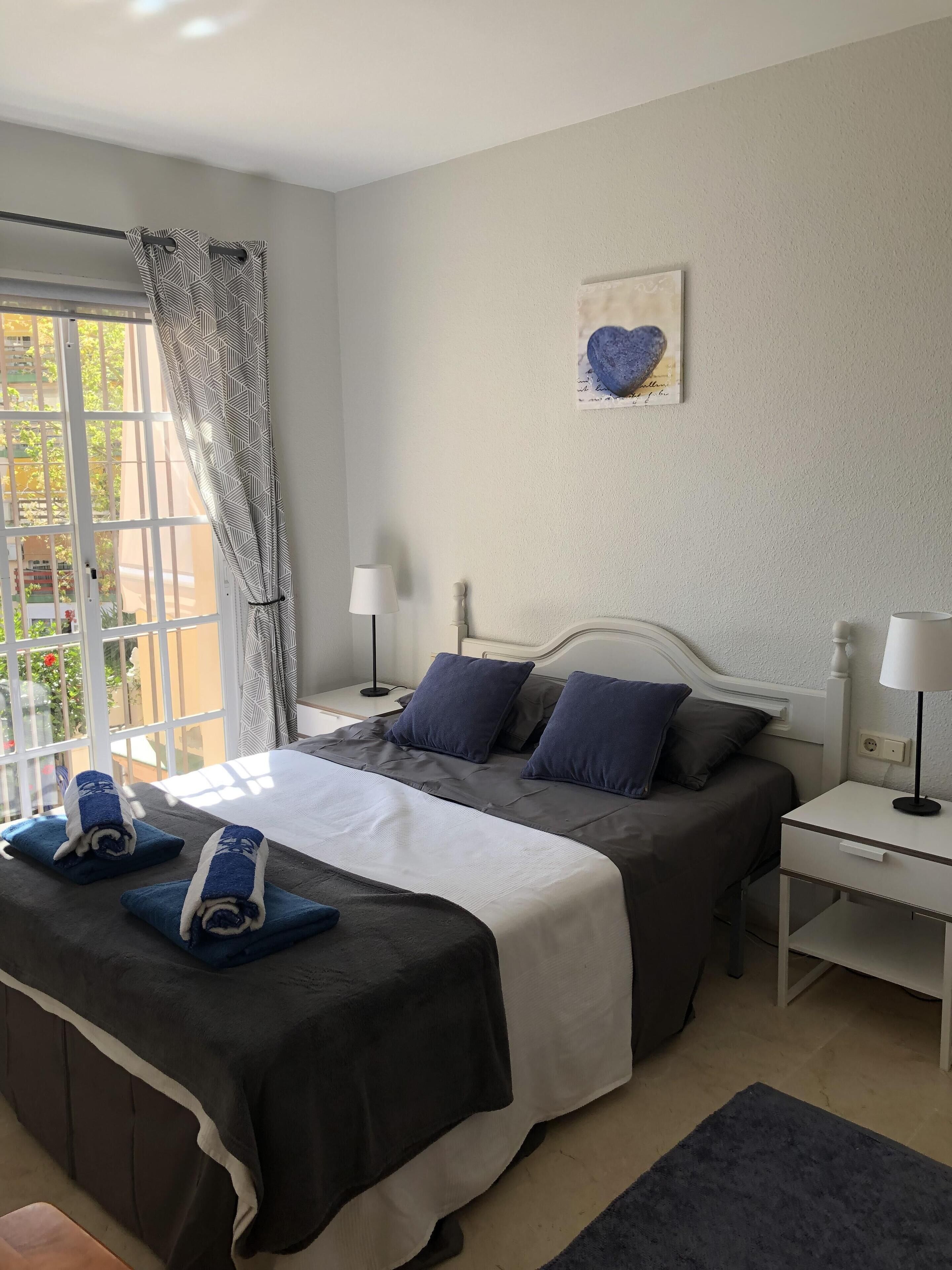 A bedroom with a bed, two bedside tables, a window with curtains, and a wall-mounted heart-shaped decoration.