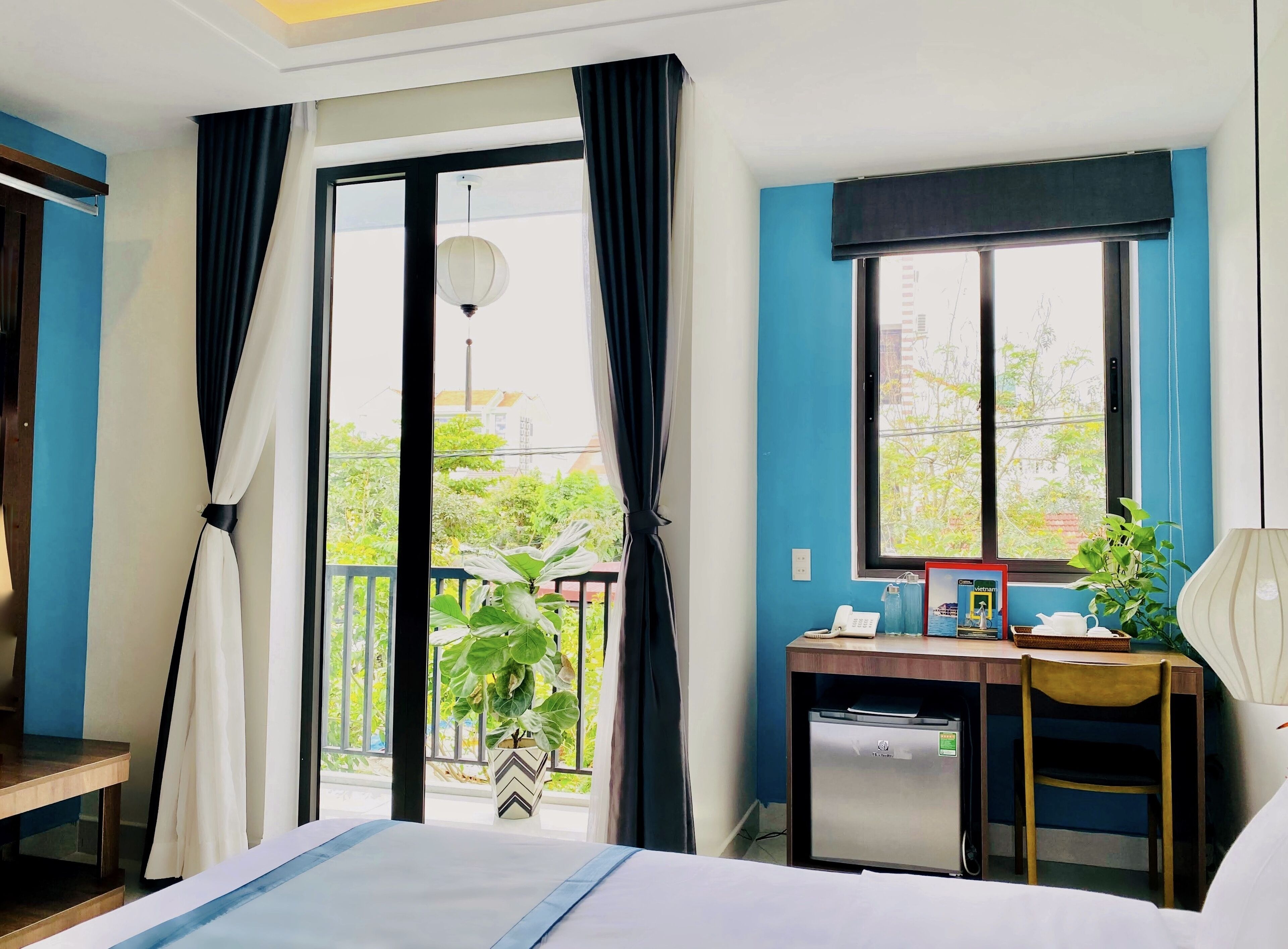 Quarto de hotel com cama, escrivaninha com computador, cadeira, pequeno frigorífico e janela com cortinas.
