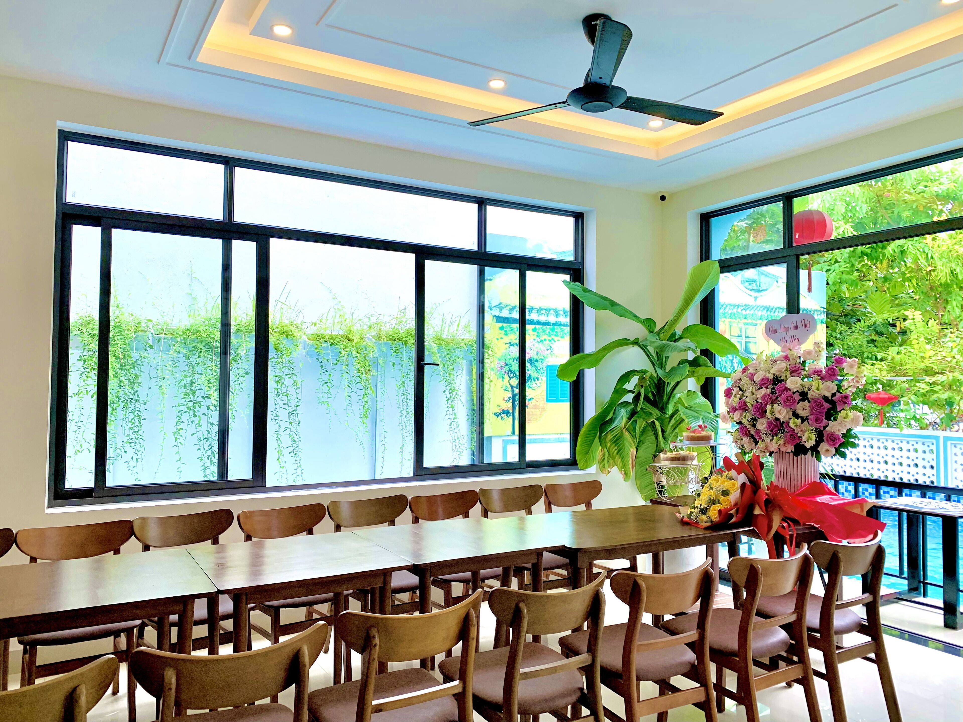A dining area with a long table and multiple chairs, large windows with greenery outside, a ceiling fan, and a potted plant with flowers.