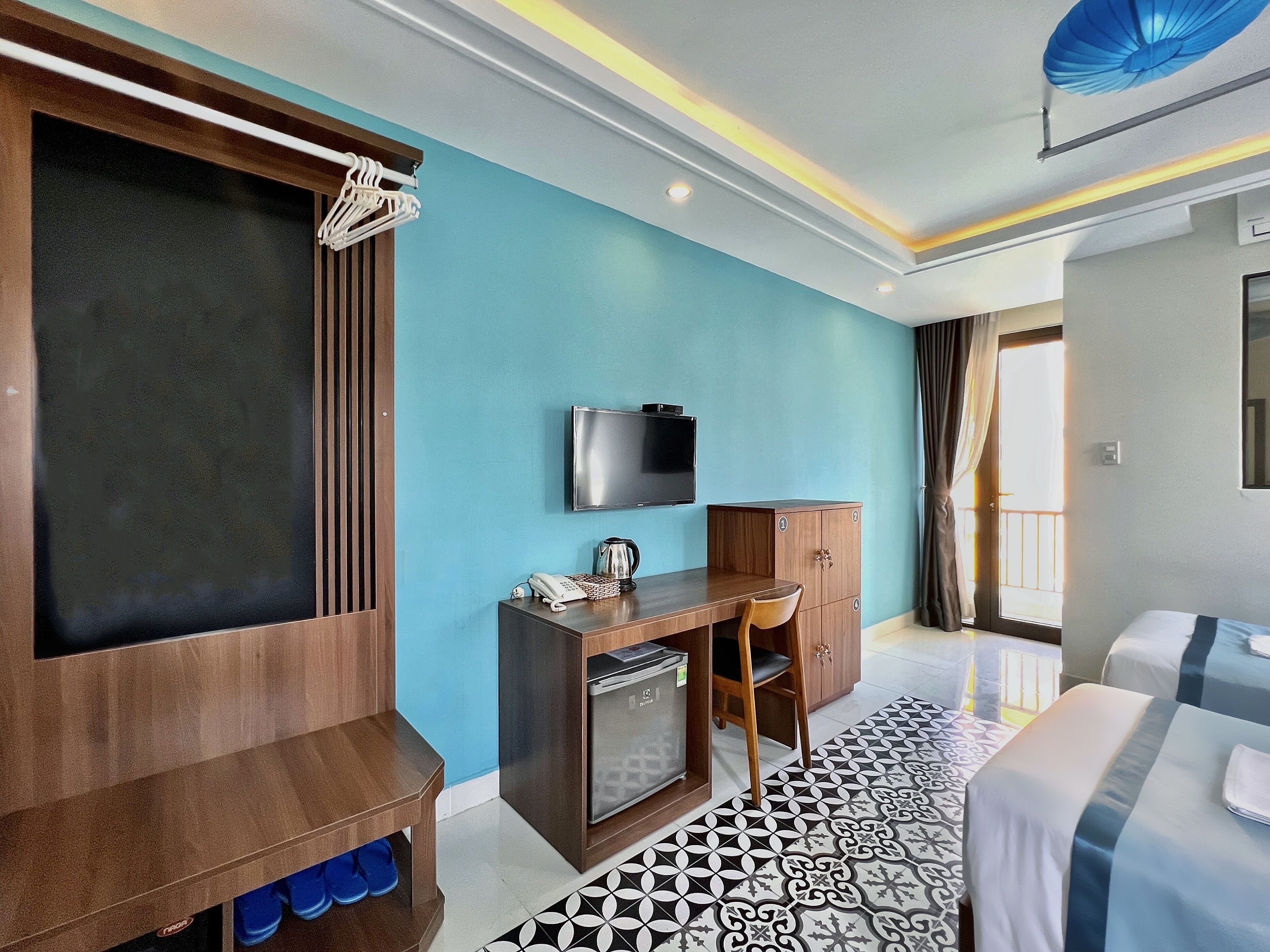 A hotel room with a wooden desk, a television, a minibar, a bed, and a patterned floor.