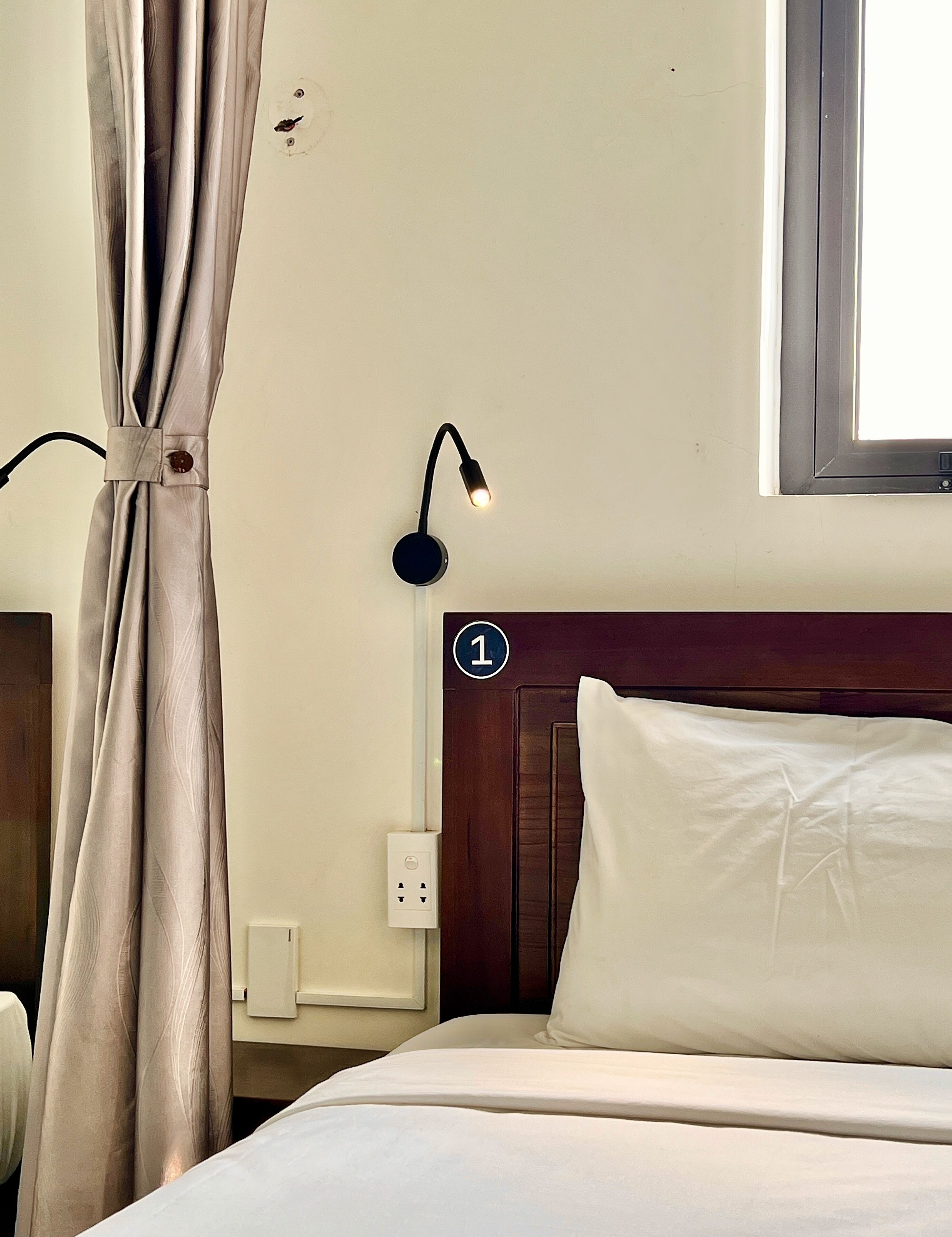 Een hotelkamer met een bed, nachtkastje, een aan de muur bevestigde lamp, gordijn en raam.