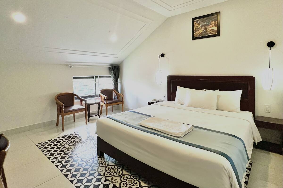 A hotel room with a bed, a desk, two chairs, a painting on the wall, and a patterned rug.
