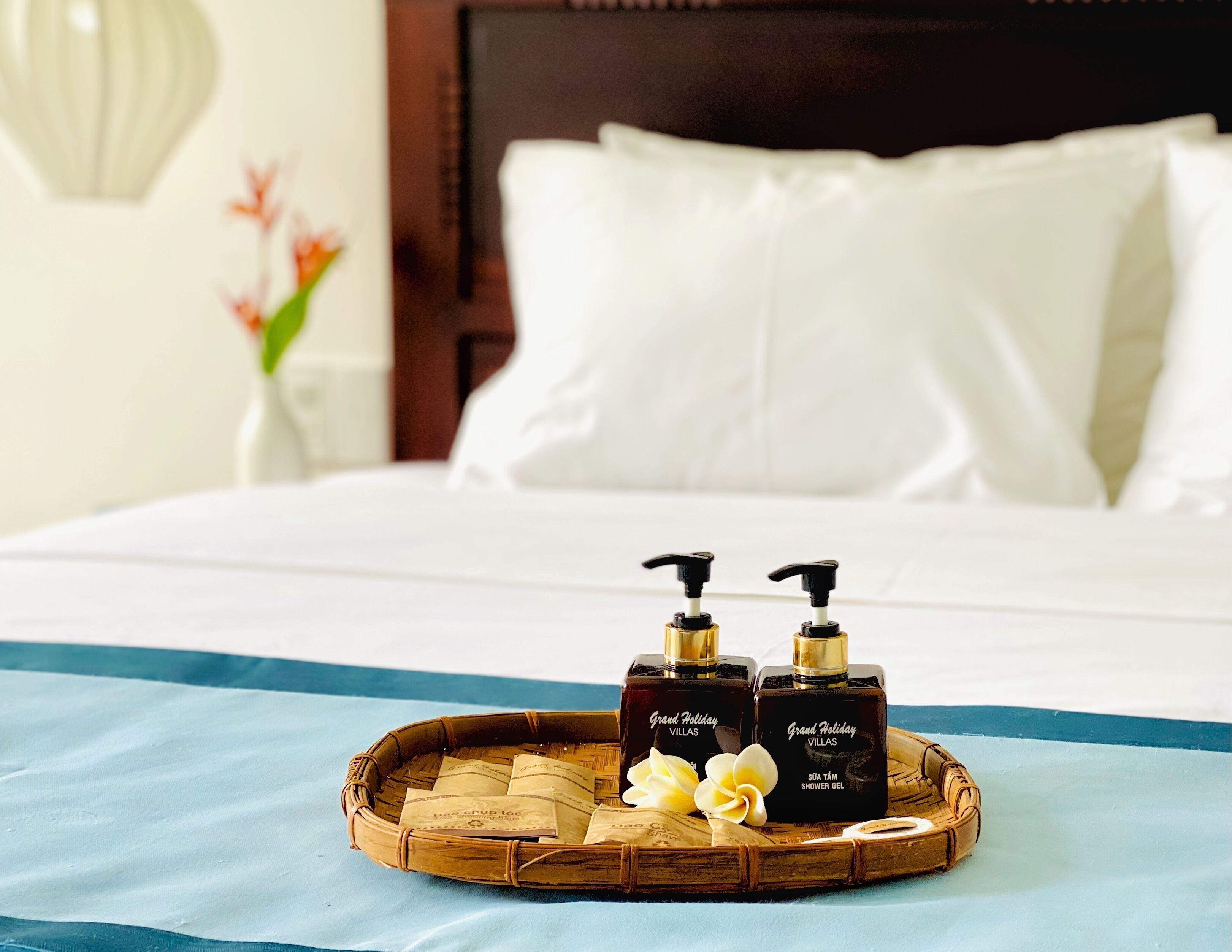 A hotel room with a bed, white pillows, a blue bedspread, and a tray with two bottles of shower gel and a flower.