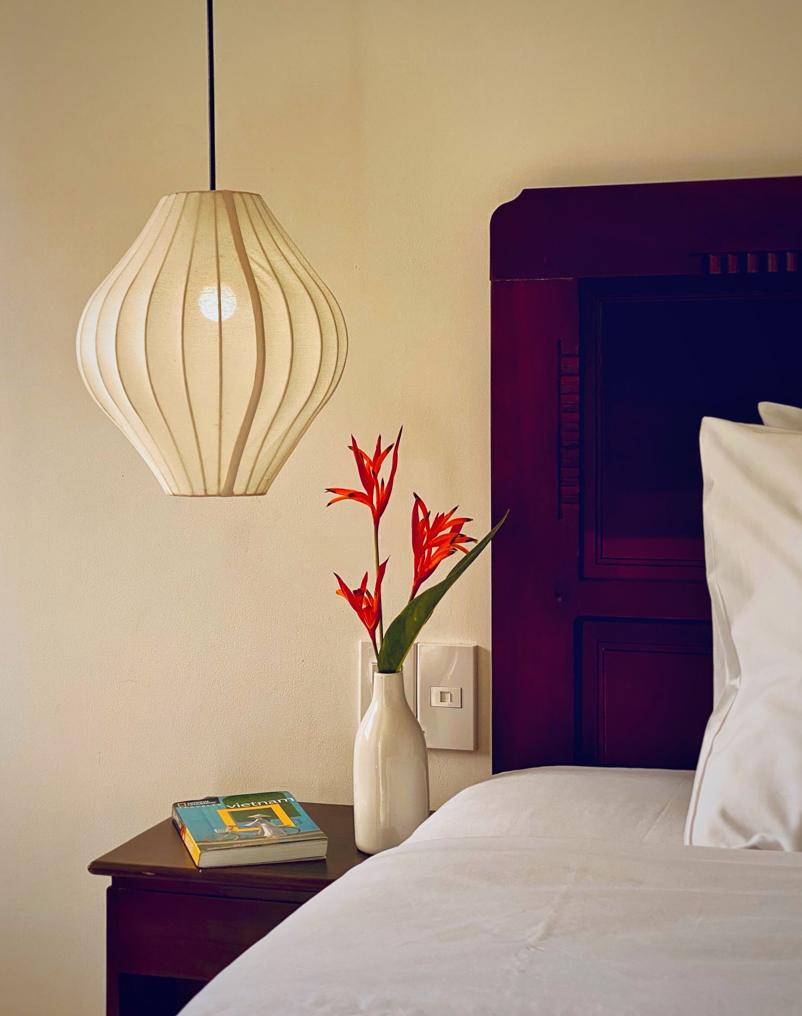 A hotel room with a bed, a nightstand with a book, a vase with flowers, a wall-mounted lamp, and a door.