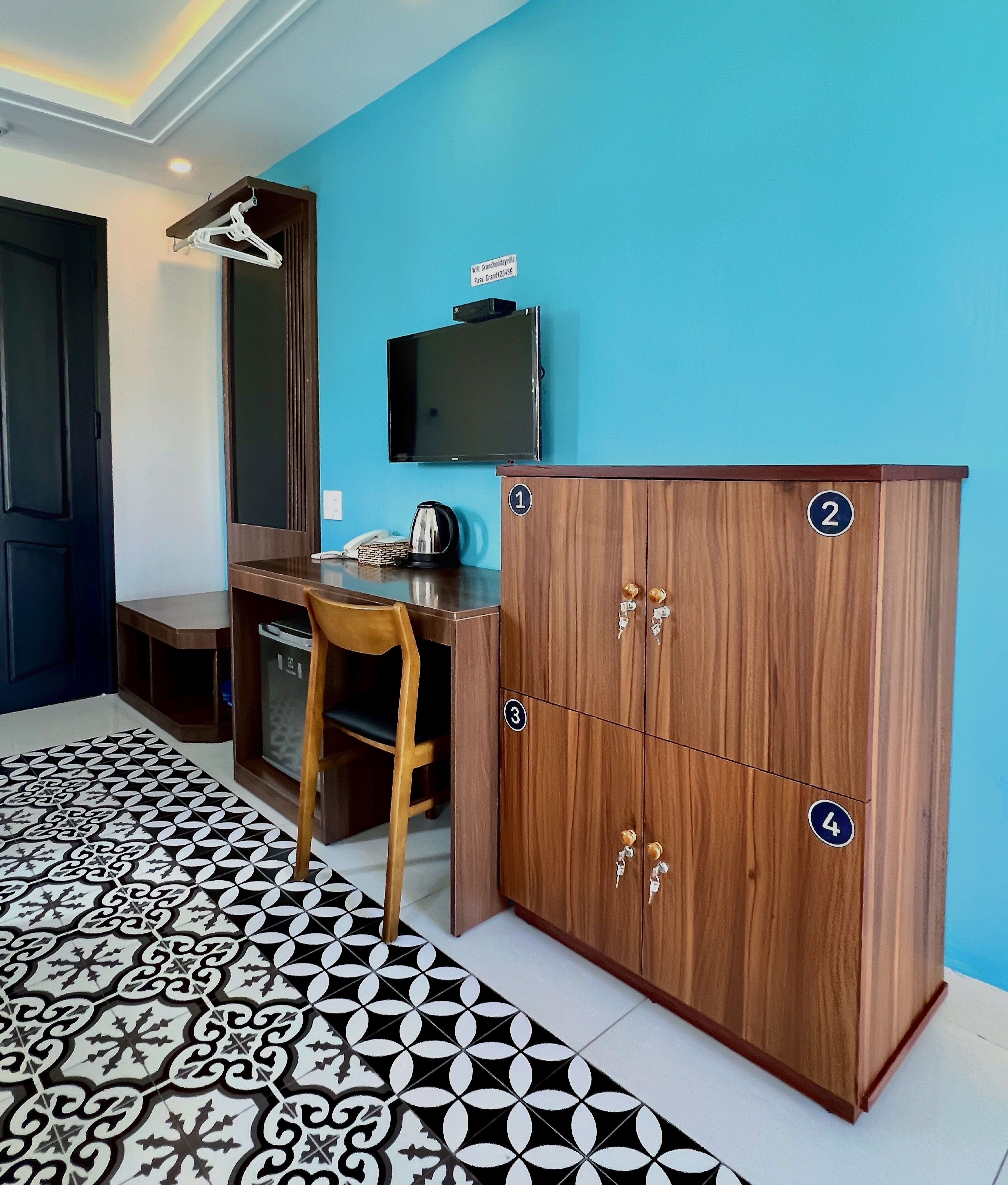 A room with a wooden cabinet numbered 1-4, a desk with a chair, a television mounted on the wall, and patterned floor tiles.
