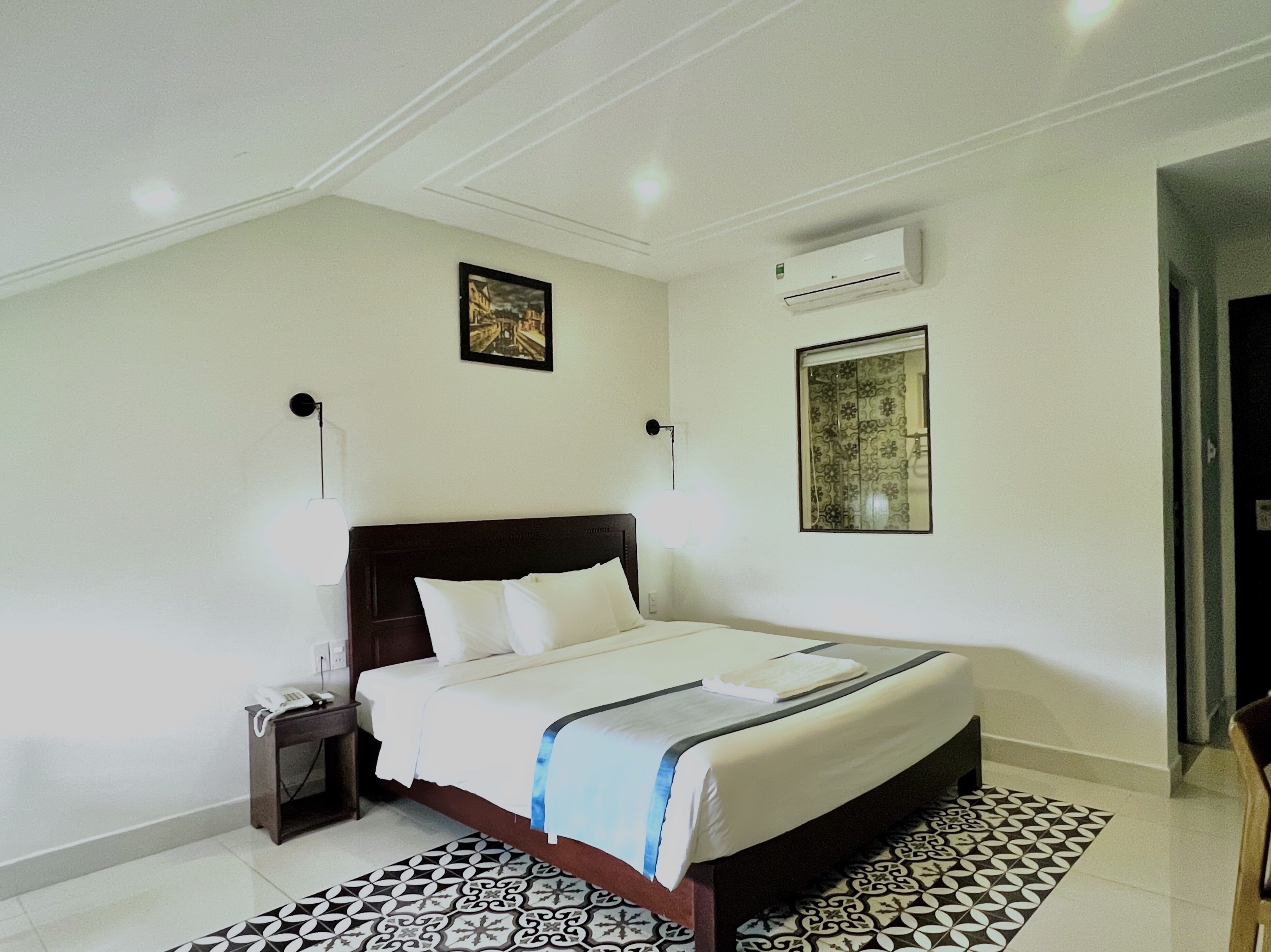 A hotel room with a large bed, a bedside table with a lamp, an air conditioning unit, and a patterned floor.