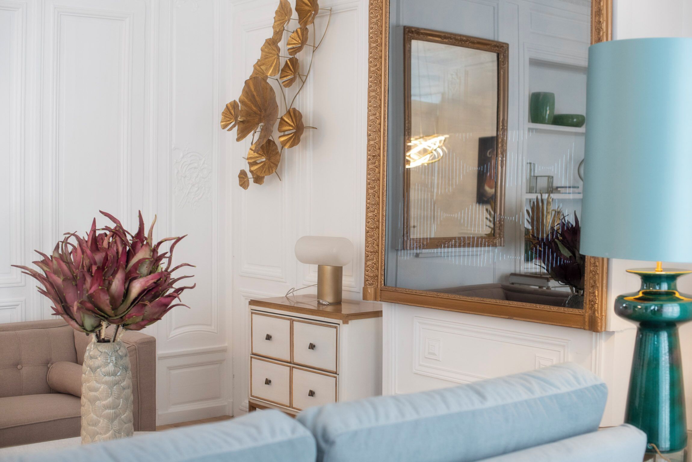 Un salon avec un canapé beige, une table d’appoint blanche surmontée d’une lampe, et un miroir doté d’un cadre doré.