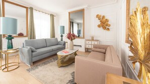 A modern living room with a grey sofa, beige armchair, and a round coffee table. There are two lamps, a mirror, and decorative wall art. - Beaubois, 2BR/3BA, 6 people (París)