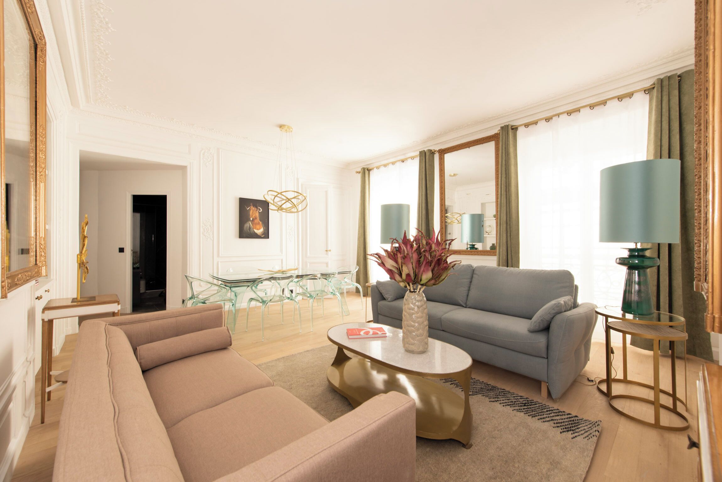 A spacious living room with a beige sofa, a grey couch, a coffee table, and a vase with flowers. There is a glass table with chairs, a mirror, and a lamp.