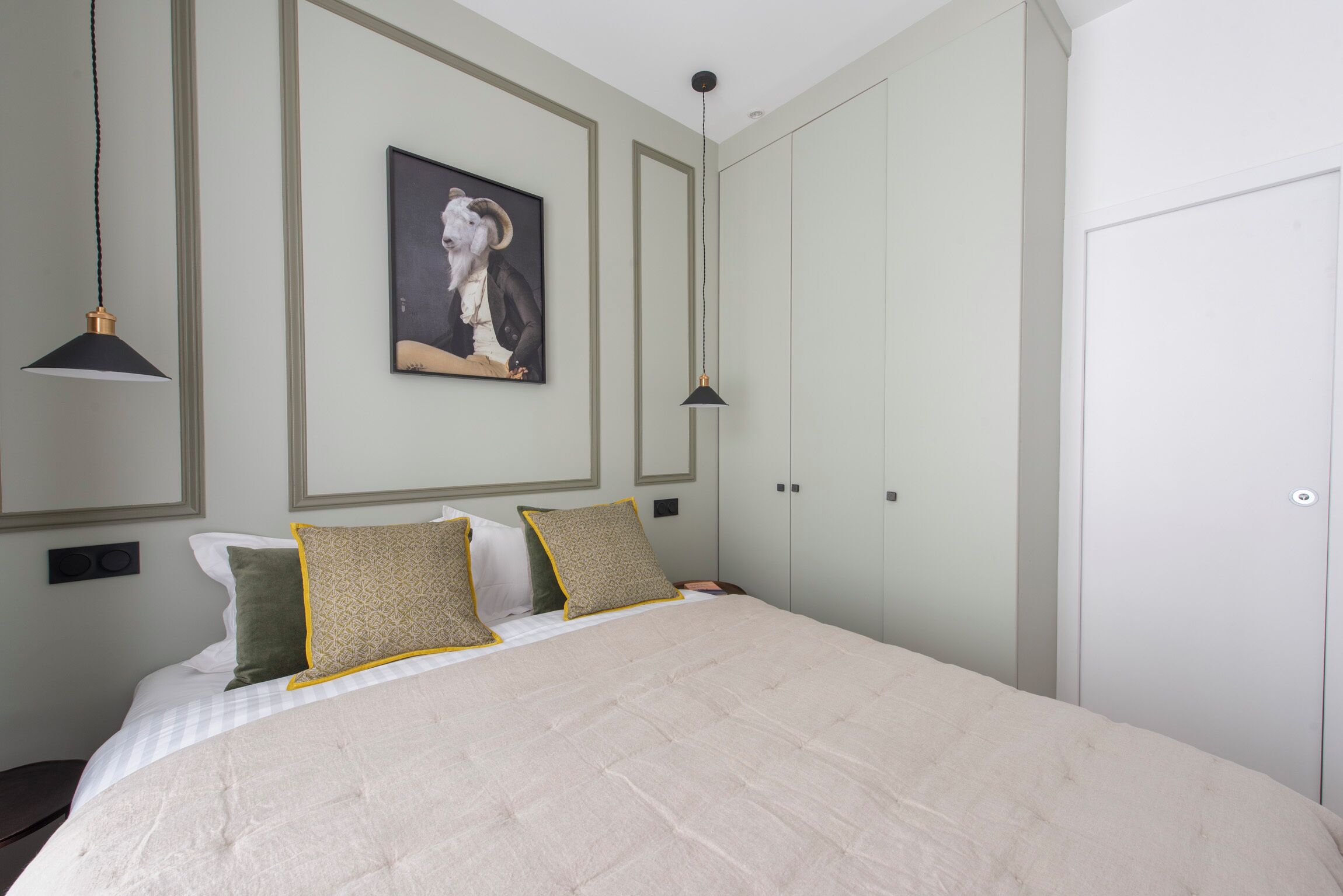 A neatly made bed with a quilted cover, two bedside lamps, a framed picture on the wall, and a wardrobe with a door.