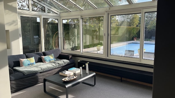 A sunlit room with a large window overlooking a swimming pool, a sofa with cushions, and a coffee table.