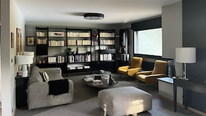 A modern living room with a sofa, ottoman, and yellow armchair, a bookshelf, and a large window.