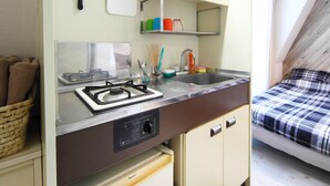 Una kitchenette compacta con estufa, lavabo y refrigerador, una ventana pequeña y una cama con una colcha a cuadros.