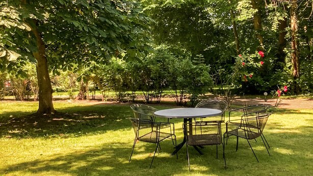 Mesa redonda con cuatro sillas en un área de jardín sombreada, rodeada de árboles.