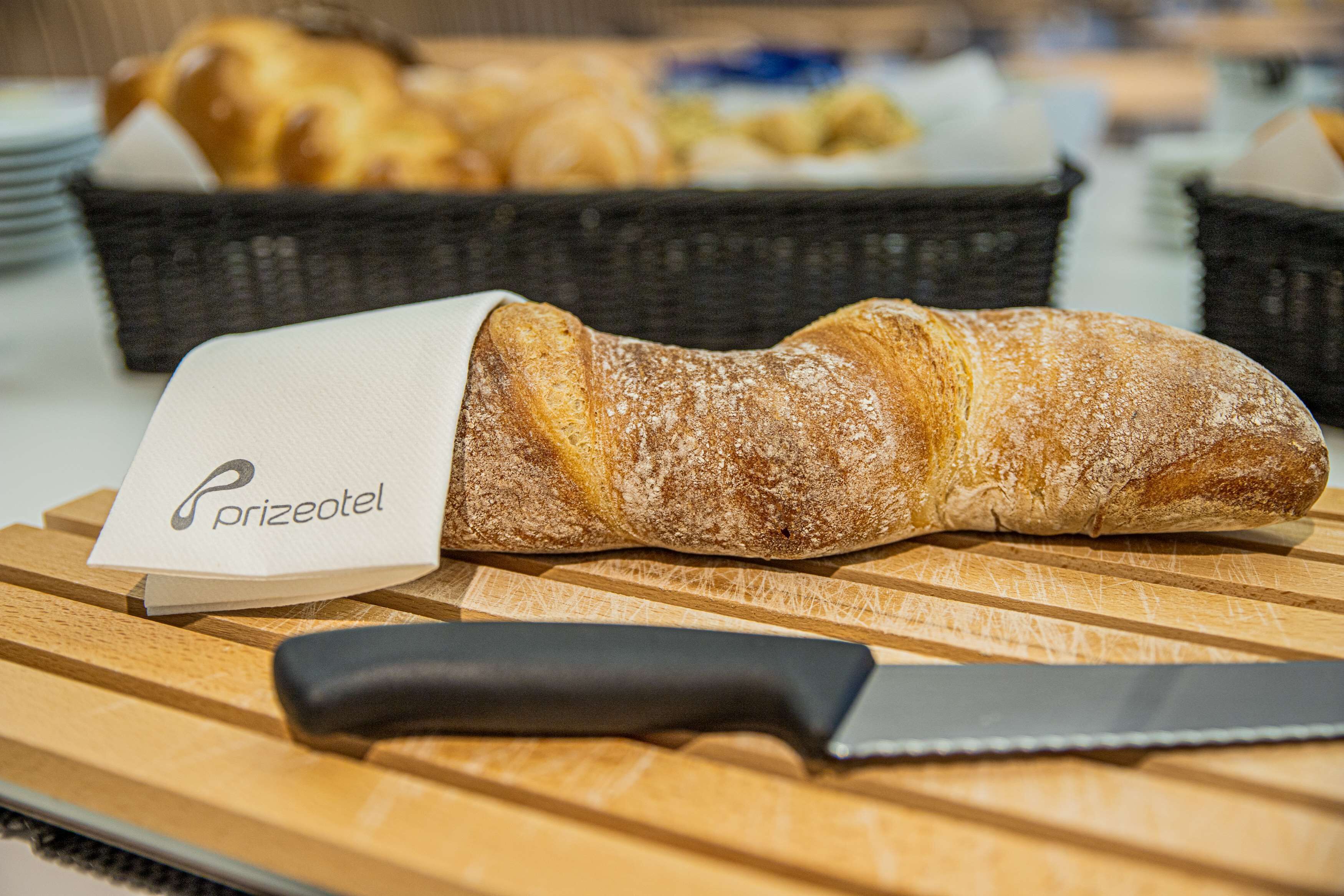 Uma barra de pão sobre uma tábua de corte de madeira, com uma faca e um guardanapo com a inscrição "prizeotel" em primeiro plano.