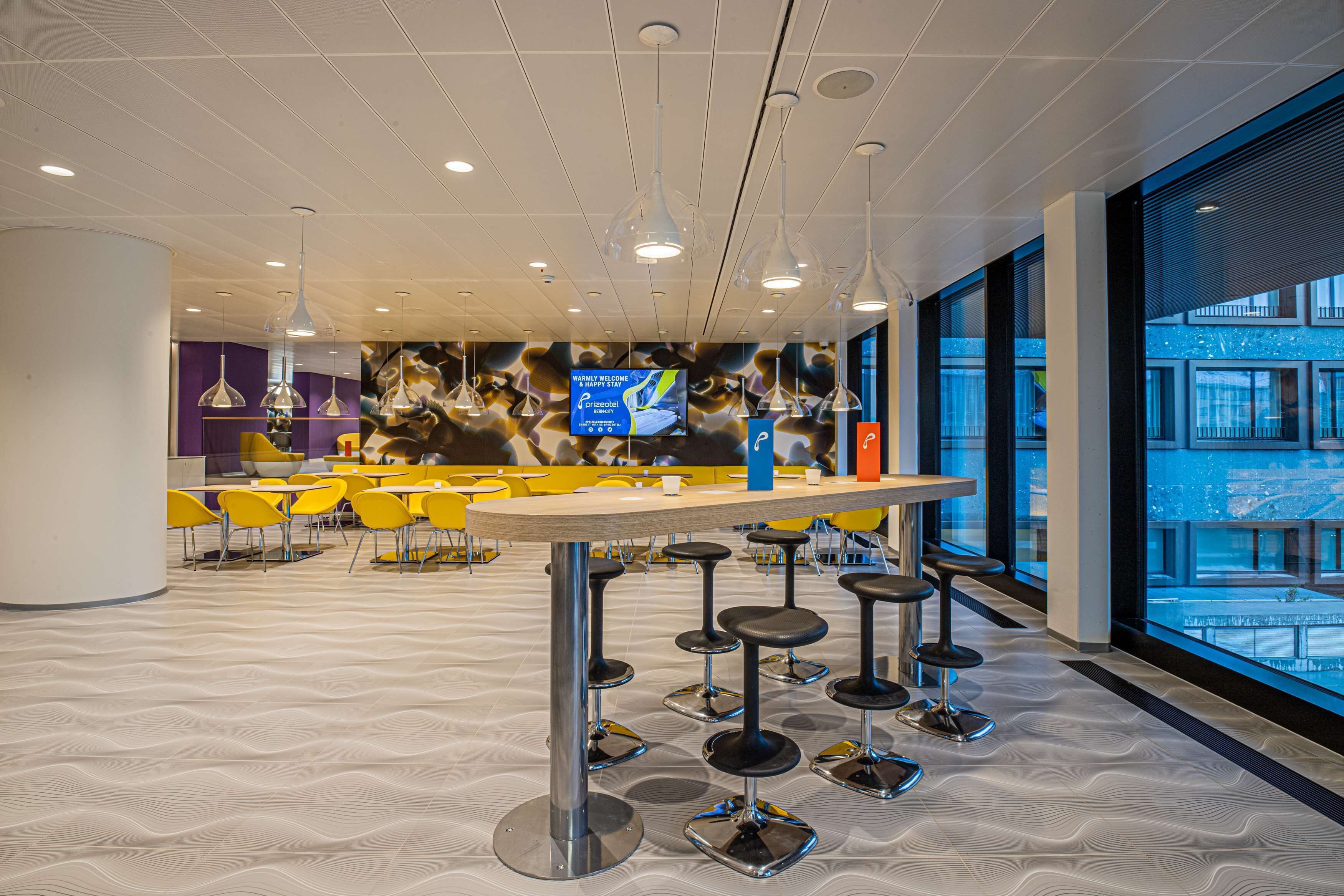 Un espace bar moderne avec une table centrale, de nombreux tabourets et un grand écran diffusant une publicité.