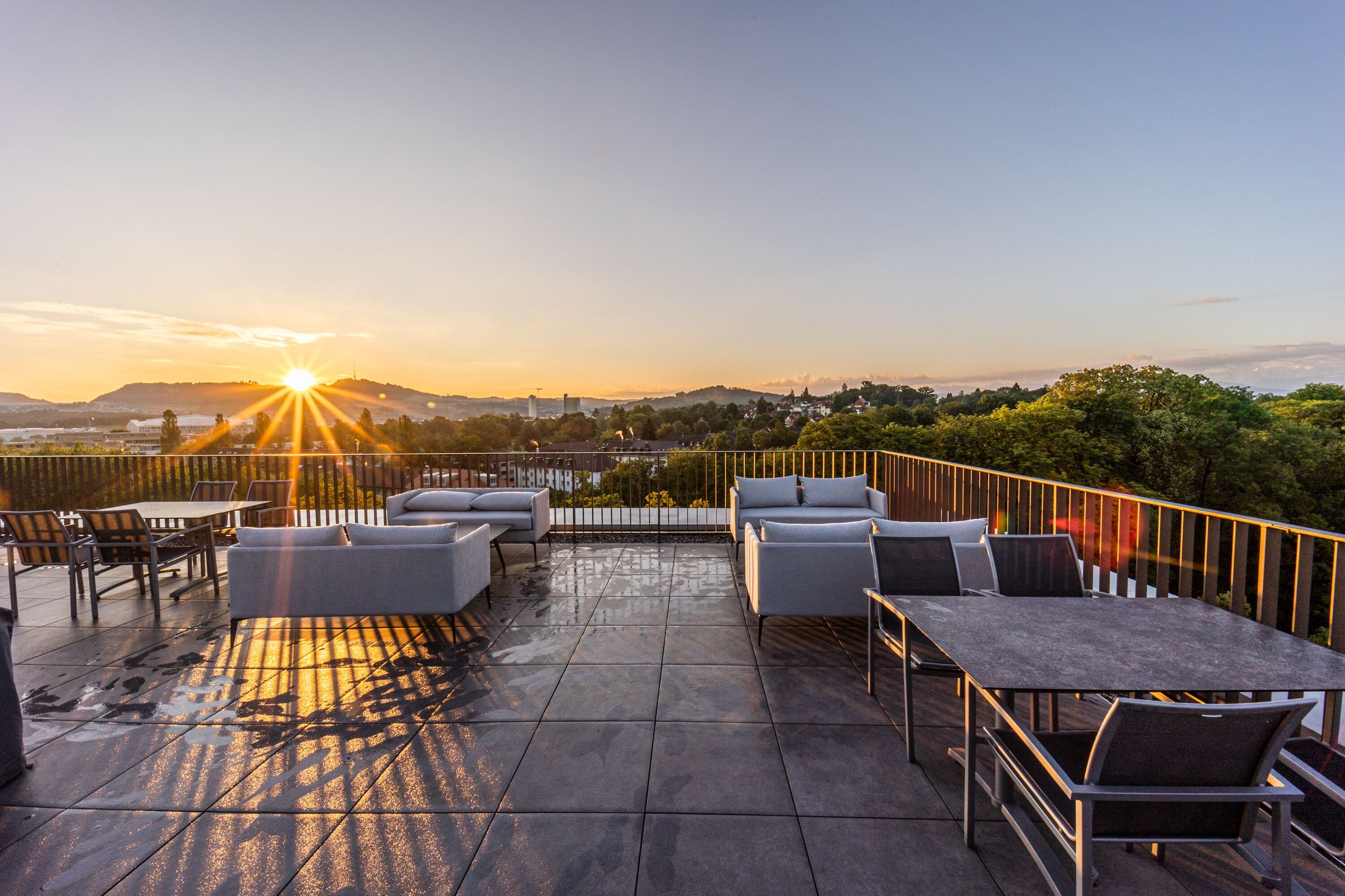 A rooftop terrace with outdoor seating, a view of the cityscape, and a sunset.