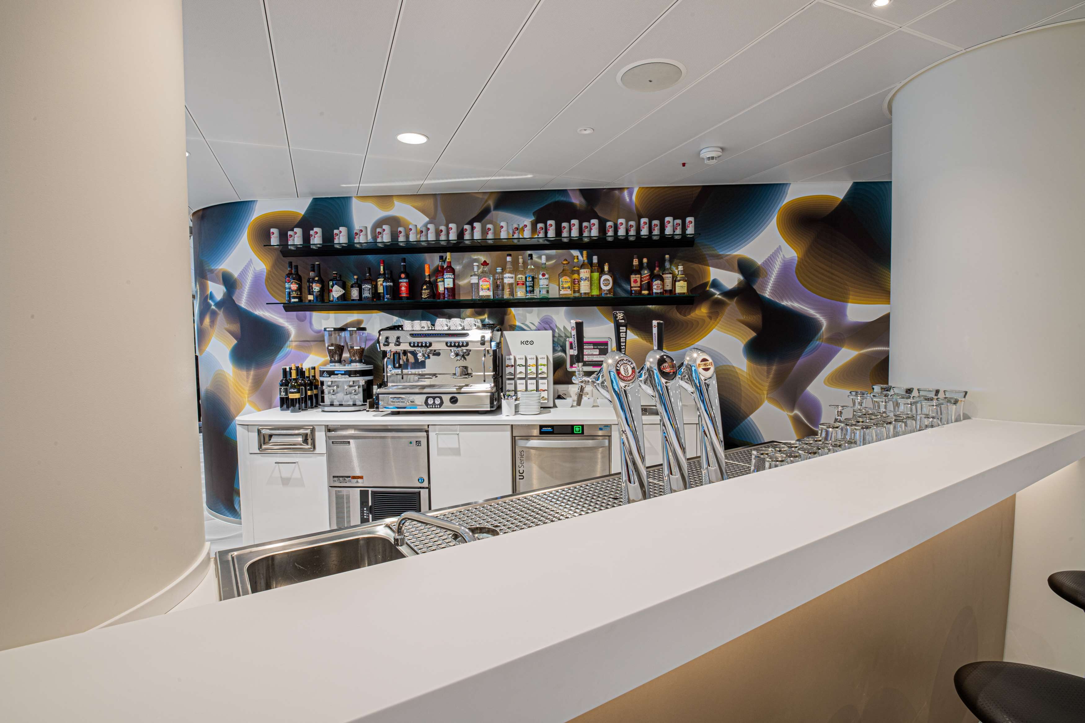 A modern bar area with a white counter, shelves with various bottles, a coffee machine, and a large abstract mural on the wall.