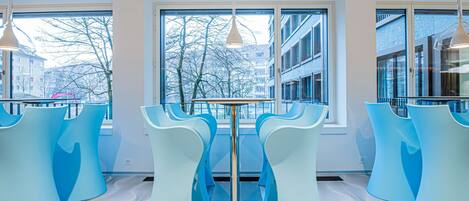 Ein modernes Interieur mit blauen Stühlen und einem zentralen Tisch, große Fenster mit Blick auf Gebäude und kahle Bäume sowie Pendelleuchten.