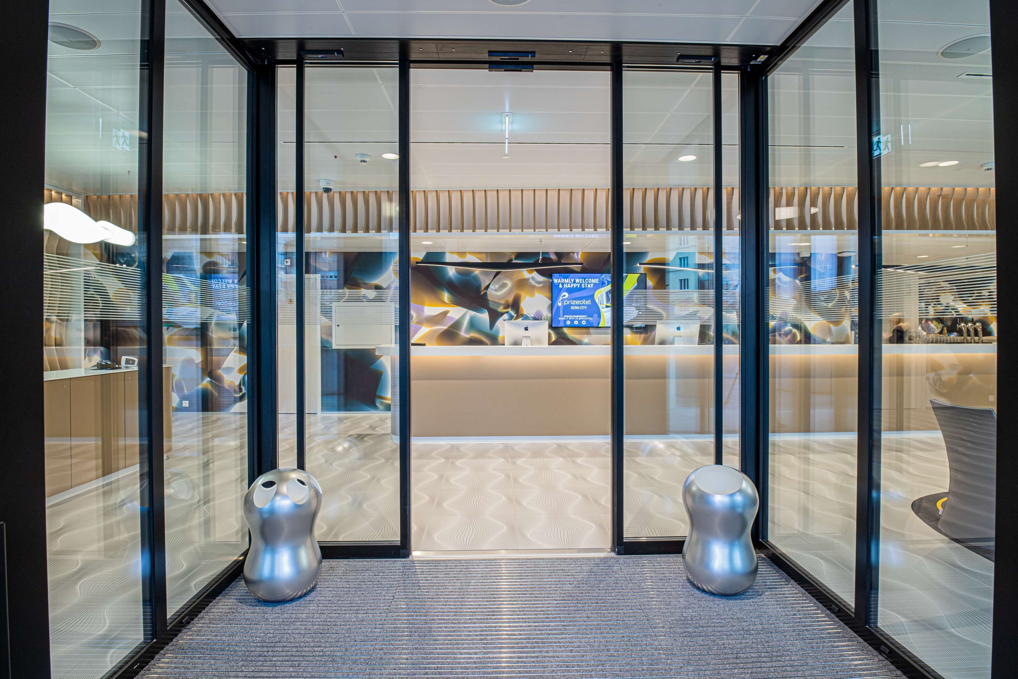 Un espace d’accueil moderne doté de portes vitrées, d’un comptoir de réception et de deux tabourets décoratifs en métal.