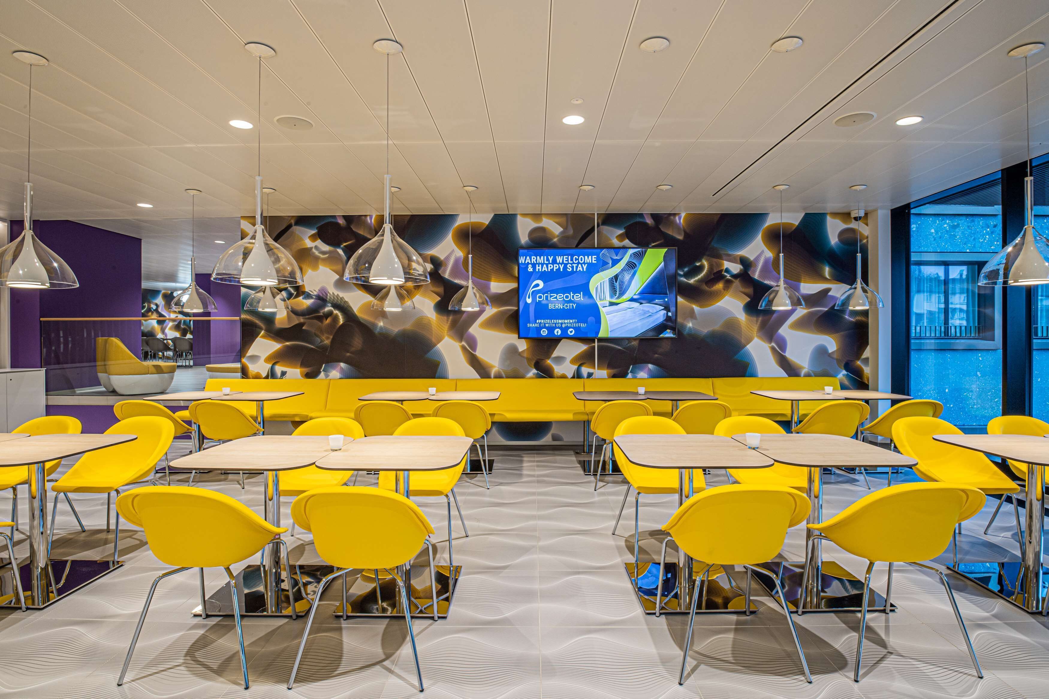 A modern dining area with yellow chairs and tables, a large digital screen displaying a welcome message, and a patterned ceiling.