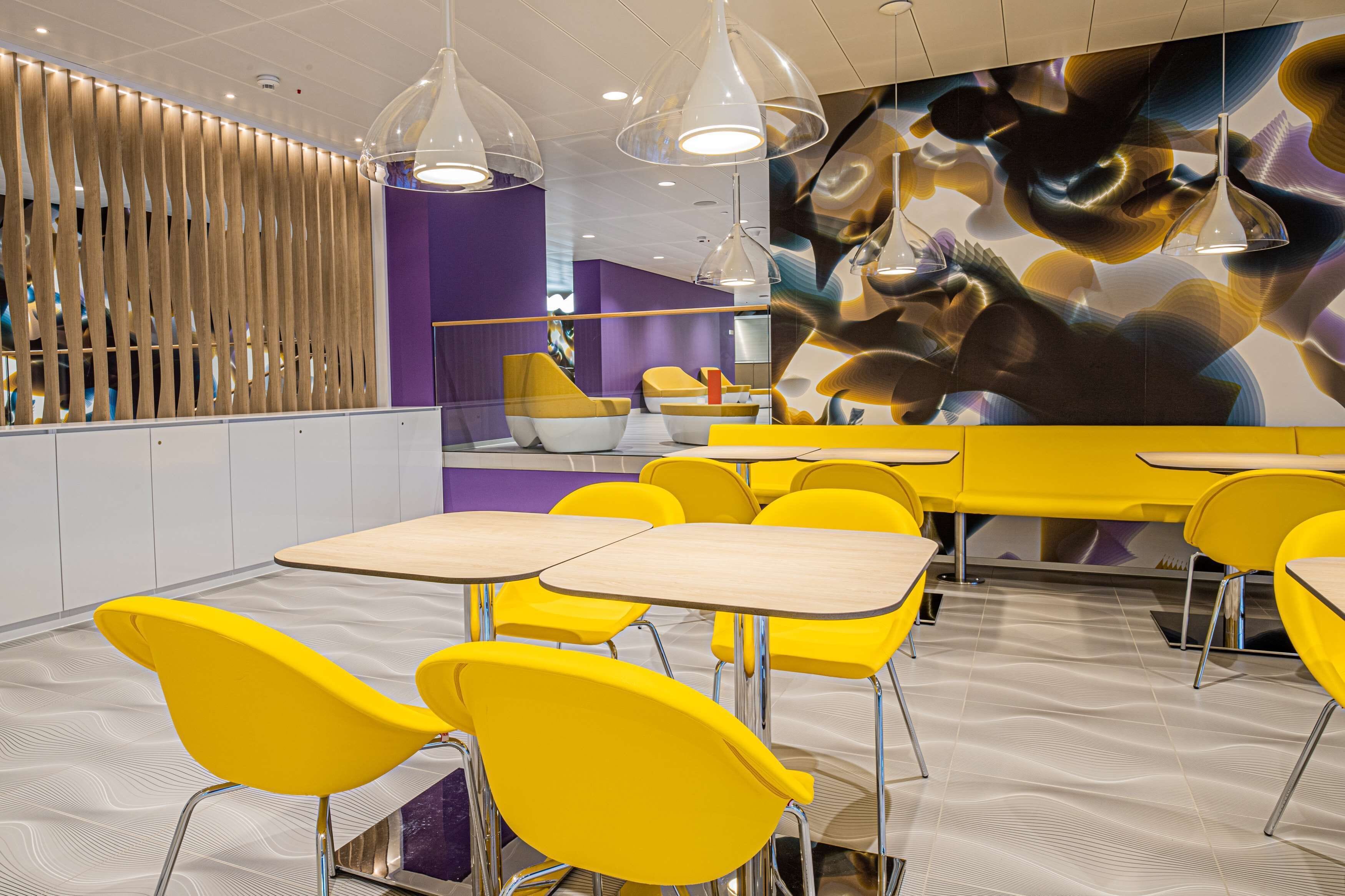 Un espace repas moderne avec des chaises et des tables jaunes, un séparateur en lattes de bois et une fresque murale abstraite et colorée.
