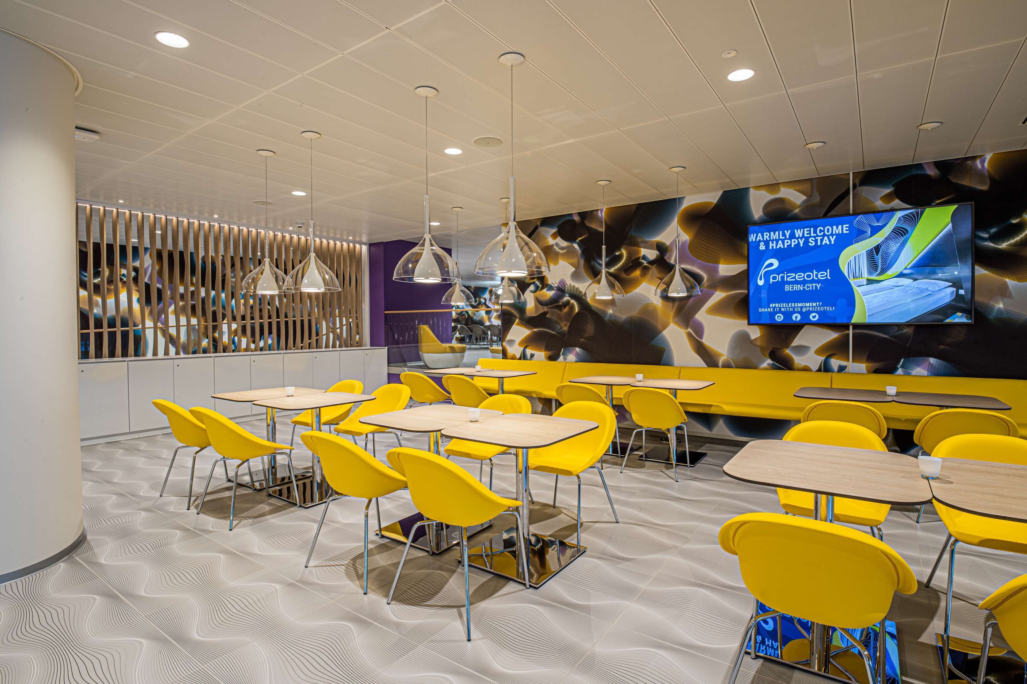 A modern hotel lobby with yellow chairs and tables, a large TV screen, and a patterned floor.
