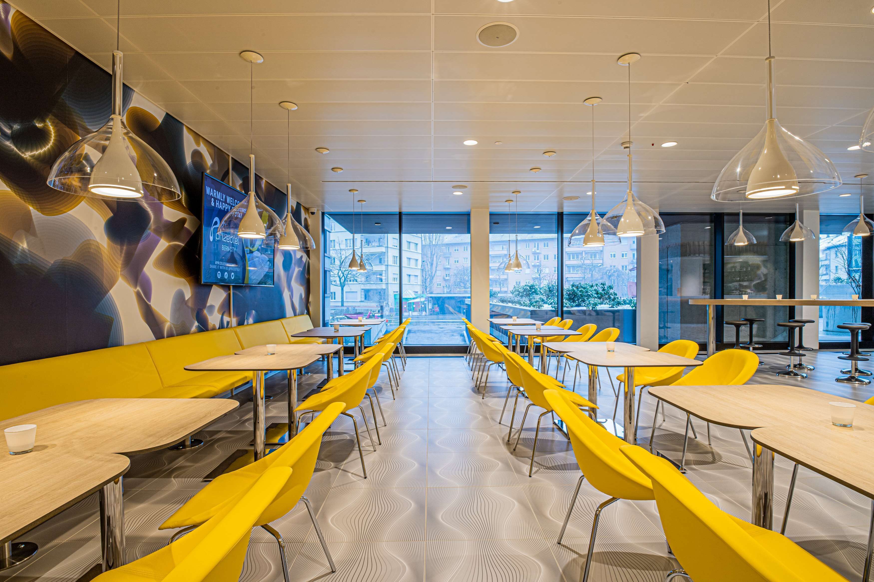 A modern dining area with yellow chairs and tables, large pendant lights, and a TV screen on the wall.