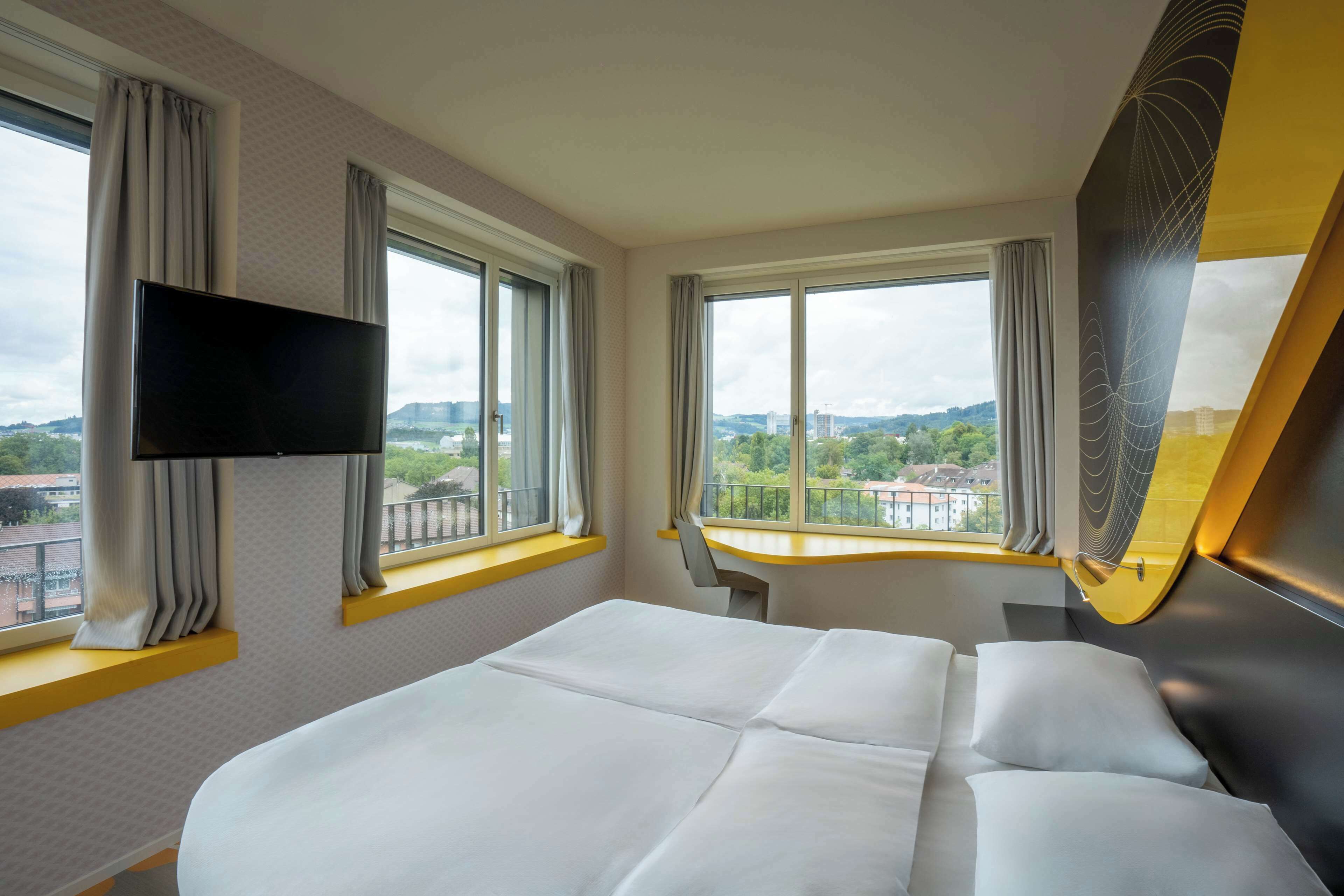Habitación de hotel con cama, televisor de pantalla plana en la pared y dos ventanas con cortinas.