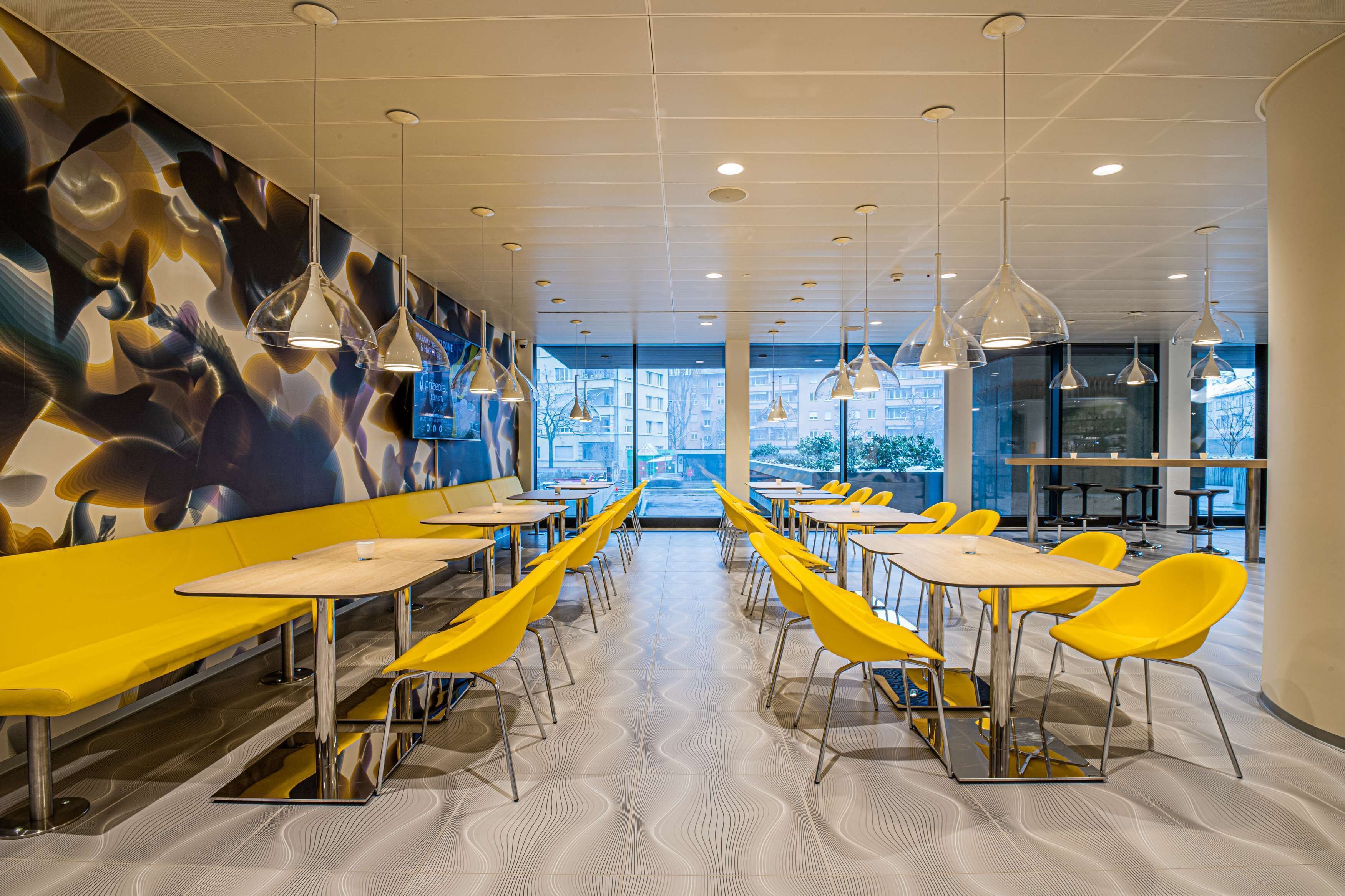 Un espace de restauration moderne avec des chaises et des tables jaunes, un sol à motifs et des luminaires suspendus.