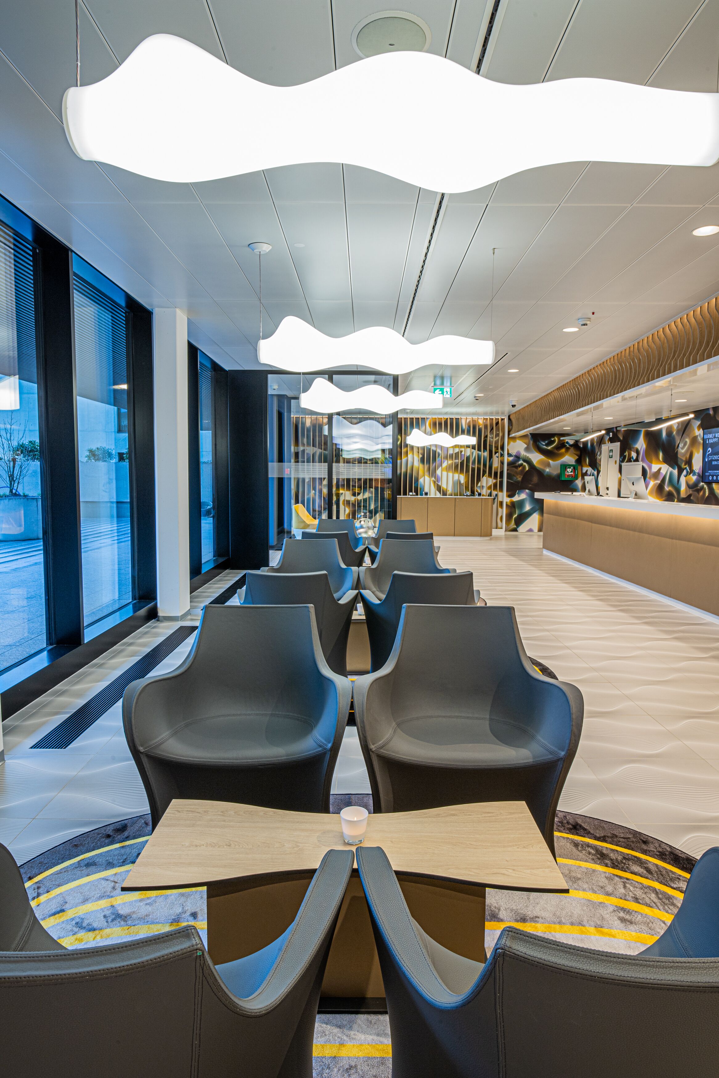 A modern hotel lobby with a reception desk, seating area with tables, and large windows offering views of the outside.