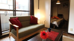 A cozy living area with a sofa, a coffee table with a vase of flowers, and a view of a balcony through a large window. - Amenity Apartment (Osaka)