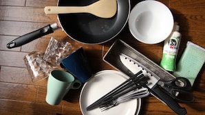 A kitchen with a frying pan, wooden spatula, white bowl, blue mug, green mug, plastic wrap, knife set, scissors, and dish soap on a wooden floor. - Amenity Apartment (Osaka)