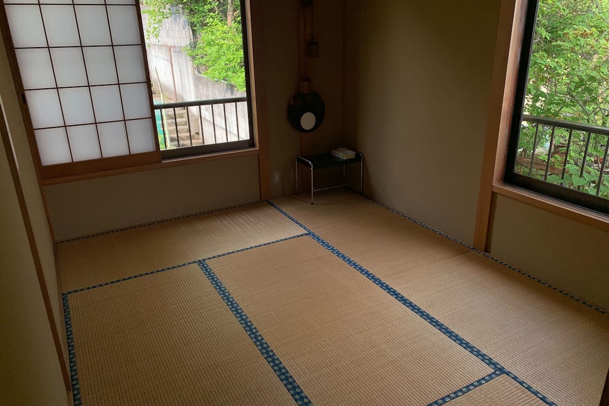 Ein Raum mit Tatami-Matten, einem Shoji-Bildschirm, einem kleinen Hocker und einer hängenden Dekoration.